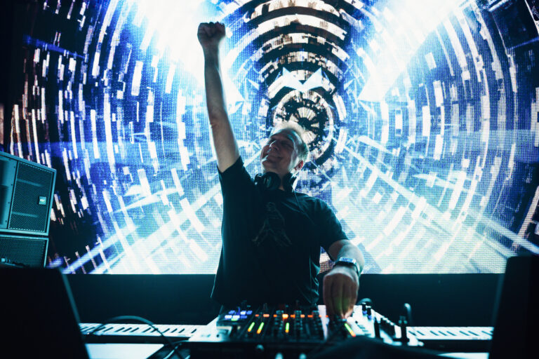 A man stands behind a mixing console in front of a large screen displaying glowing patterns, raising one arm in the air.