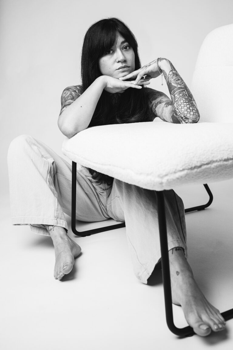 A tattooed woman with dark hair sits barefoot on the floor, resting her chin on her hands behind a white armchair in a monochrome studio portrait.