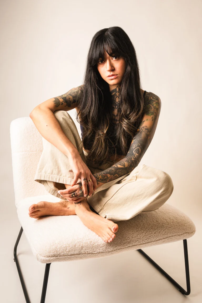A tattooed woman with long dark hair and bangs sits cross-legged barefoot in a white armchair against a plain background.