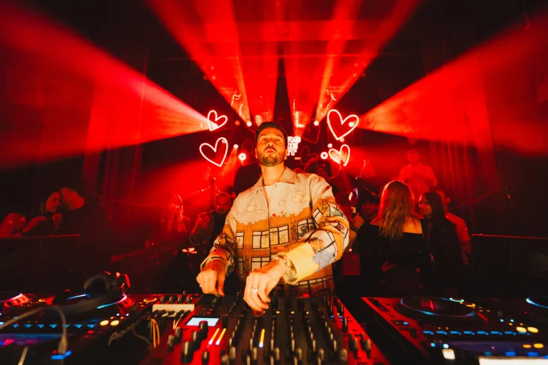 A man wearing a patterned jacket stands behind a DJ mixer, performing under intense red stage lighting and glowing heart-shaped neon signs.