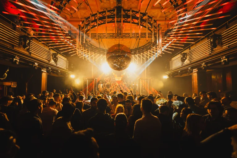 A large crowd fills a warm, orange-lit club beneath a massive circular light installation and a hanging disco ball at Kaufleuten 2025.