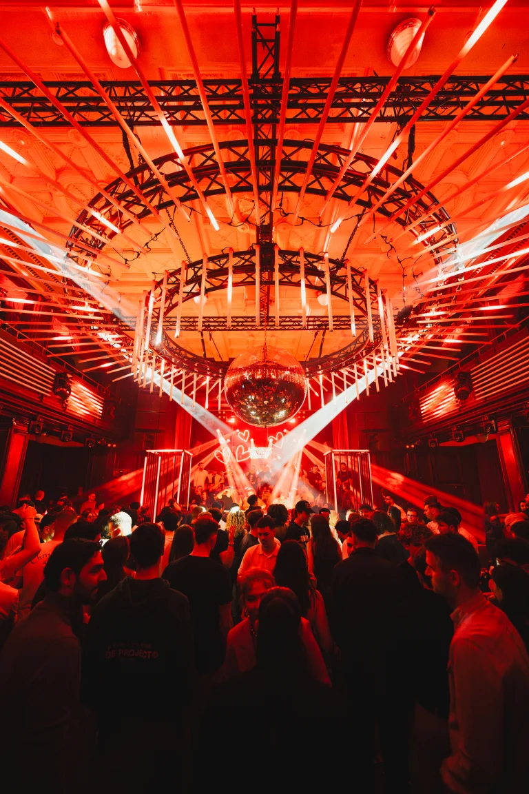A crowded nightclub lit entirely in red with a large disco ball suspended beneath an intricate overhead lighting rig.