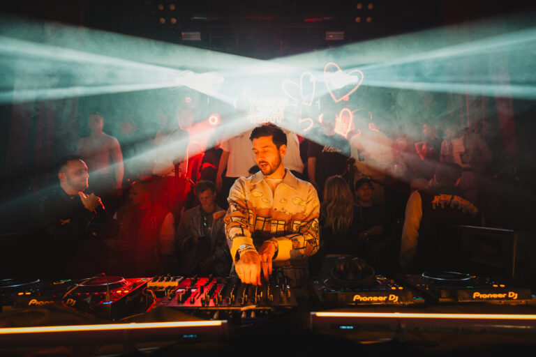 A man behind a DJ mixing console under red and white stage lights, with a crowd visible in the hazy background.