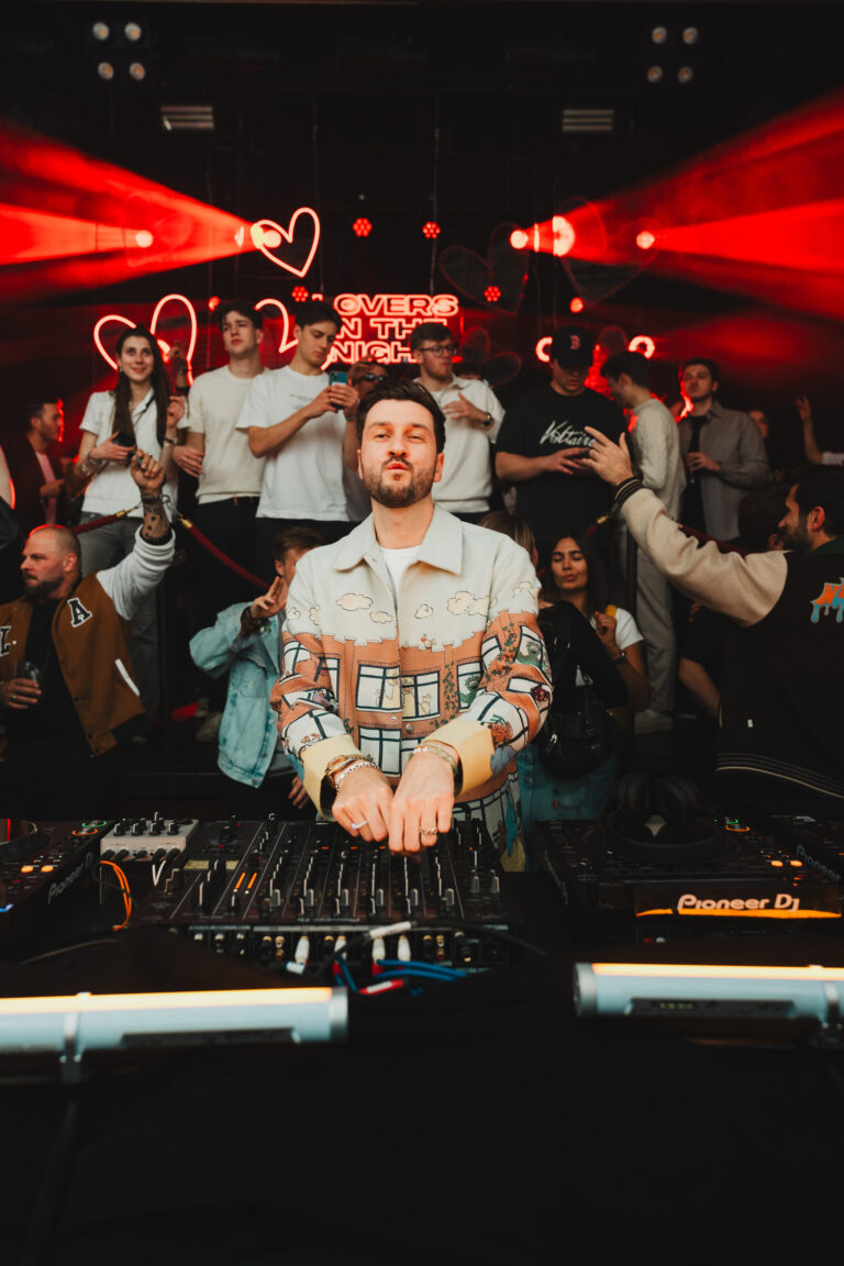 A man stands behind a DJ mixing console in a dimly lit club with a crowd and red neon signs in the background.