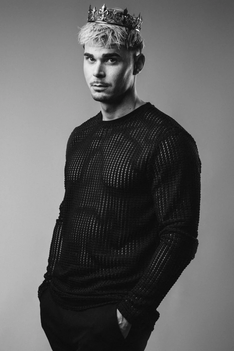 A black and white studio portrait of a man wearing a crown and a mesh sweater, looking towards the camera.