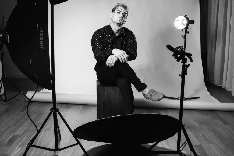 A black-and-white portrait of a man sitting on a box in a studio, surrounded by lighting equipment.