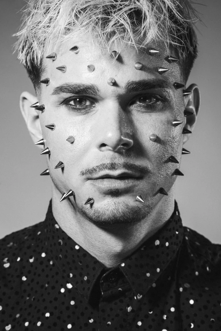 Close-up monochrome portrait of a man with small metallic spikes embedded in his face and light-dyed hair, wearing a patterned shirt.