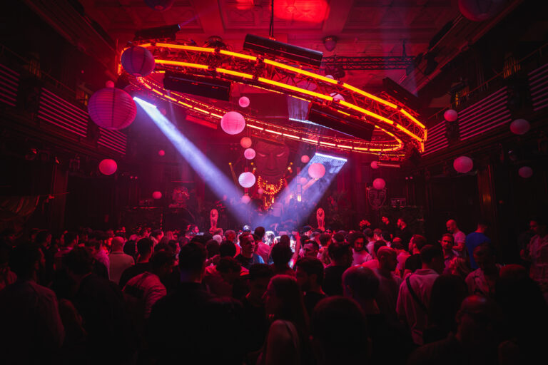 A large crowd of people gathers in a dimly lit, red-toned concert venue beneath a circular overhead rig and paper lanterns.