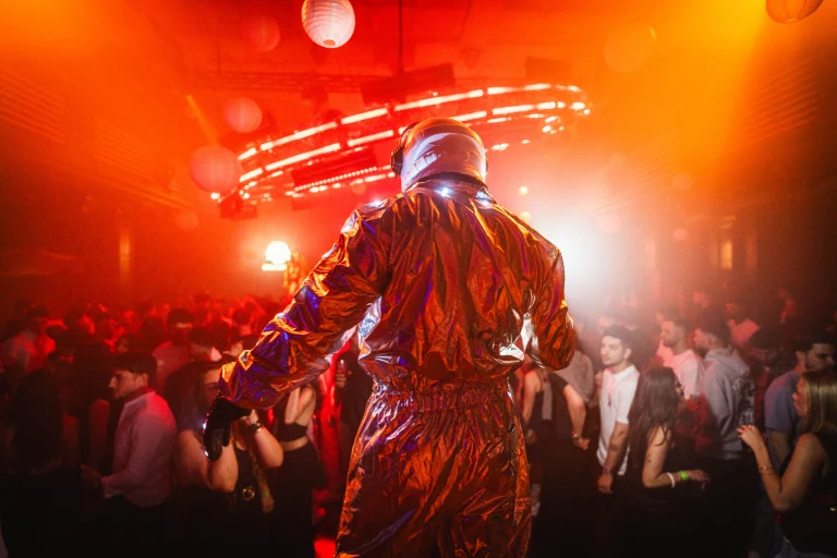 A man wearing a reflective jumpsuit and helmet faces away from the camera, overlooking a crowded indoor venue illuminated by orange stage lighting.