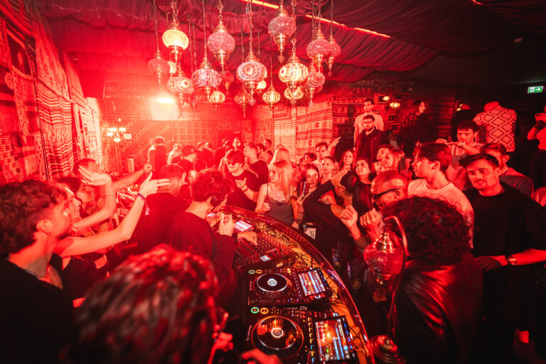A crowded nightclub lit by red ambient light, with decorative mosaic lamps hanging above a DJ booth where a man mixes music.