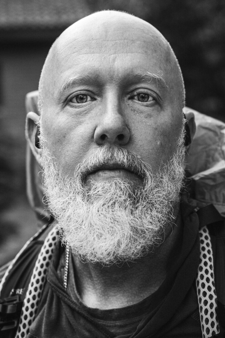 Close-up black and white portrait of a bald man with a graying beard, wearing a backpack.