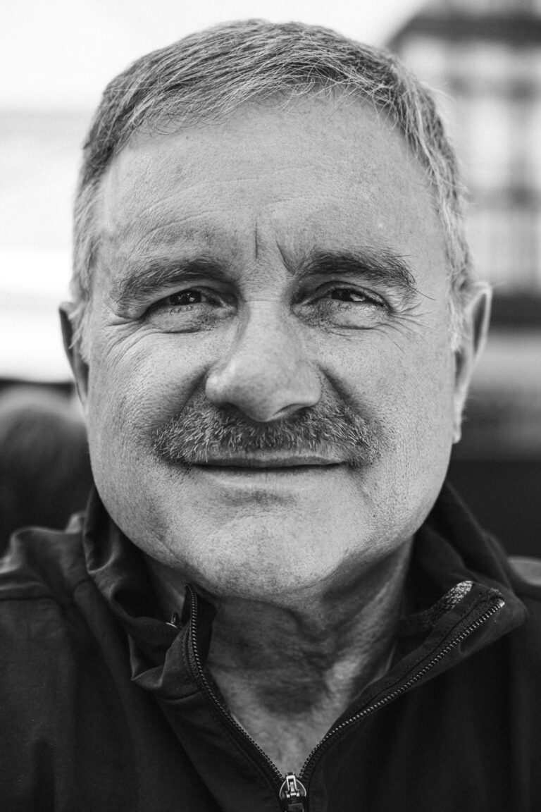 Fekete-fehér közeli portré egy mosolygó, középkorú férfiról, rövid, őszülő hajjal és bajusszal.