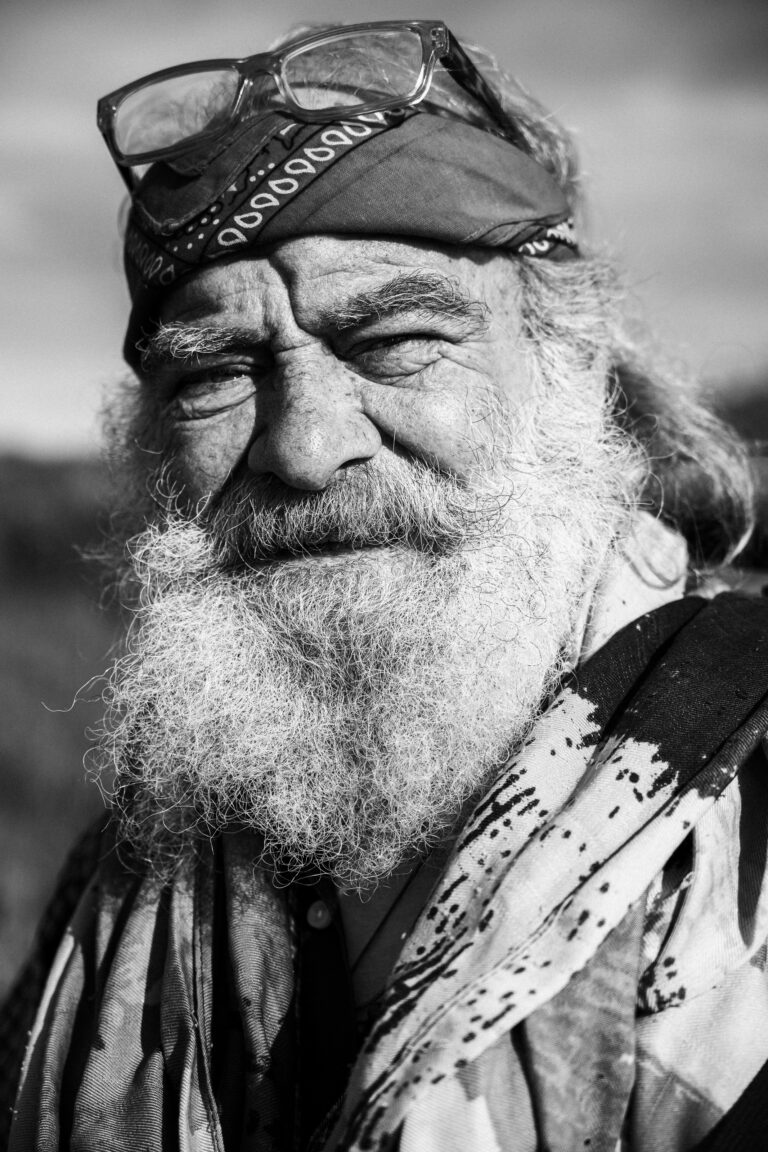 Black and white close-up portrait of a man with a long white beard, wearing a patterned bandana and glasses on his head.
