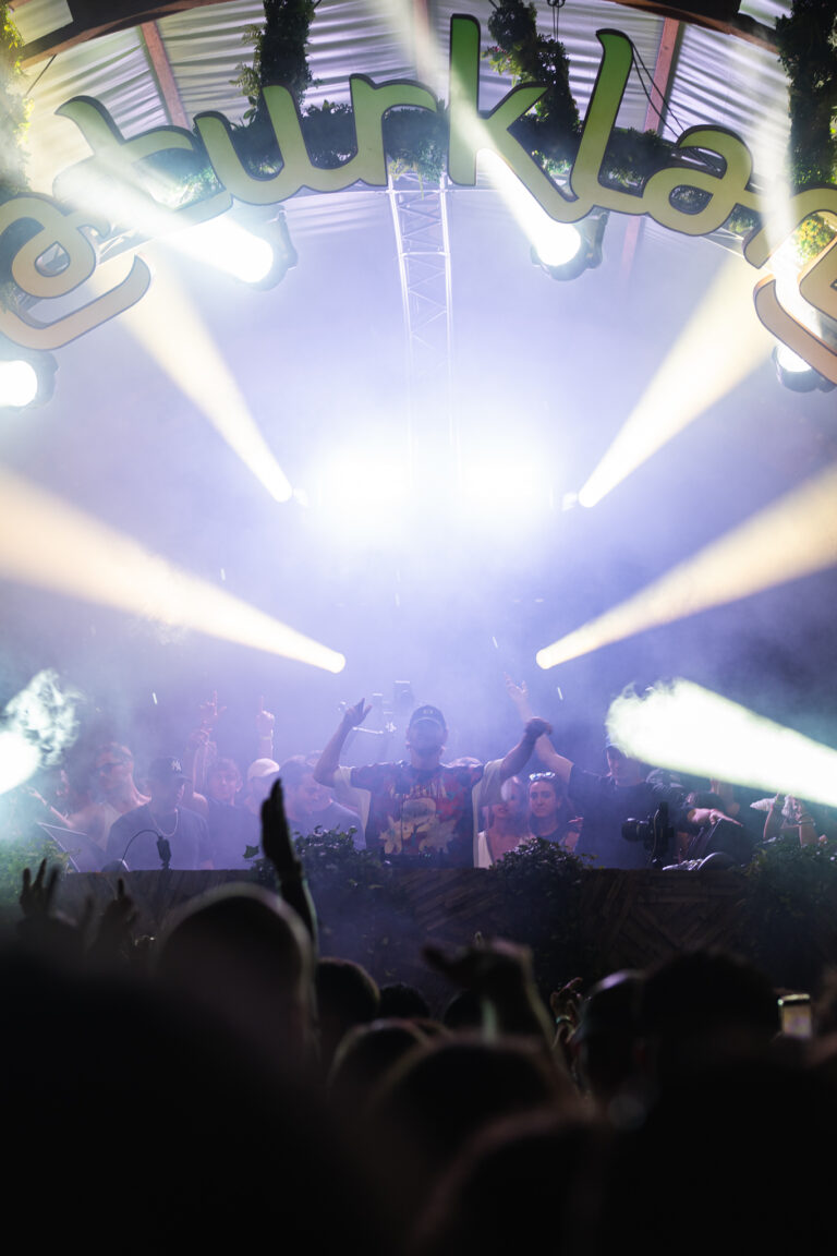 A man stands behind a stage booth with outstretched arms beneath bright stage lights and smoke, with the sign Naturklang overhead and a crowd watching below.