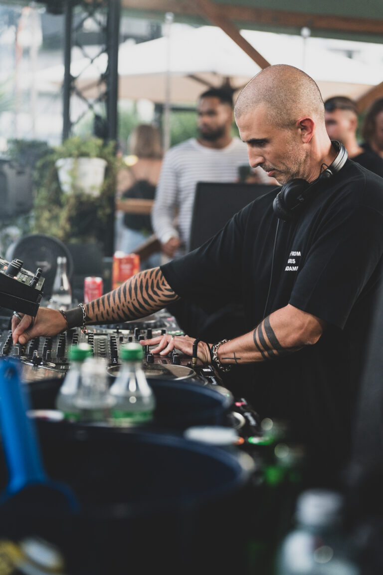 A bald man with tattooed arms wearing a black t-shirt operates a DJ mixing console outdoors.