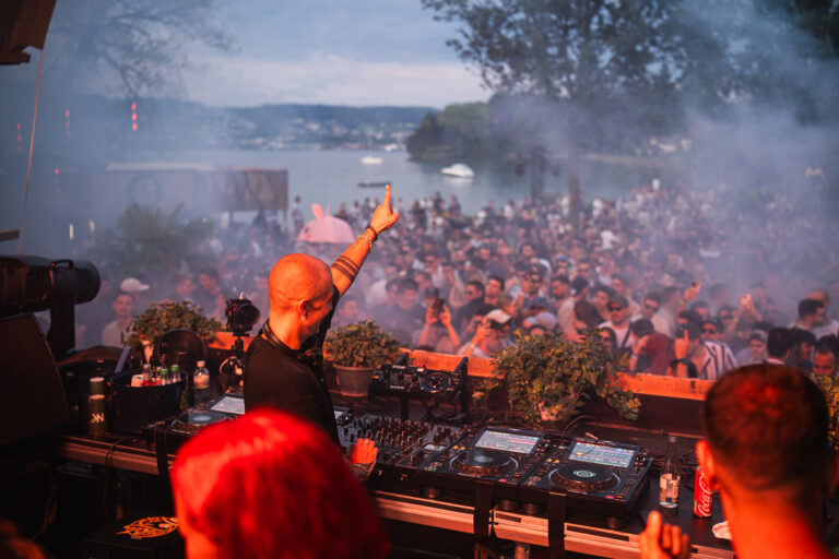 A bald man stands behind a DJ mixer on an outdoor stage, raising one finger while facing a large crowd gathered by a body of water under hazy daylight.