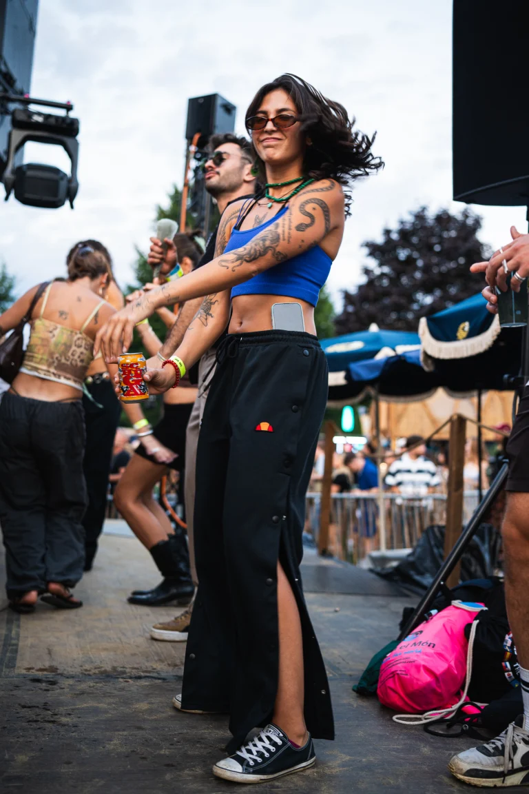 A woman wearing a blue crop top and black sweatpants dances outdoors at a festival, holding a can in her hand while a man dances behind her.