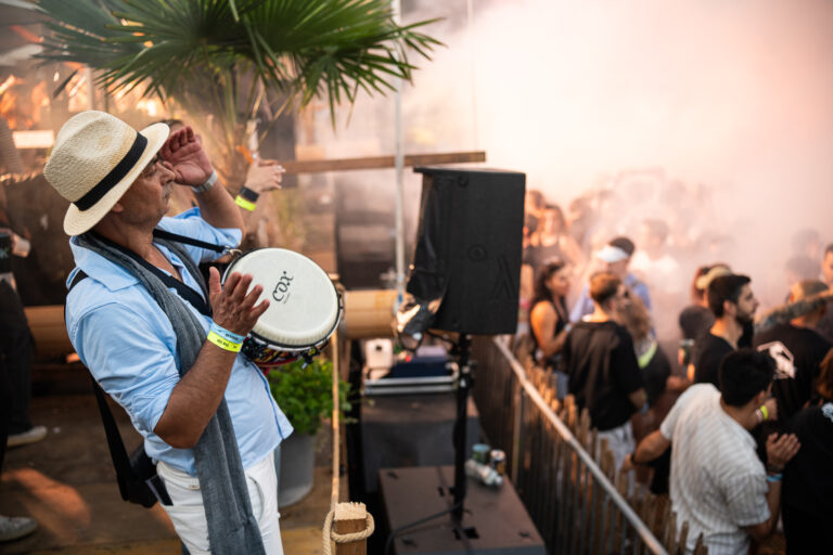 A man wearing a straw hat and a light blue shirt plays a drum outdoors near a crowd in a hazy, sunlit setting.
