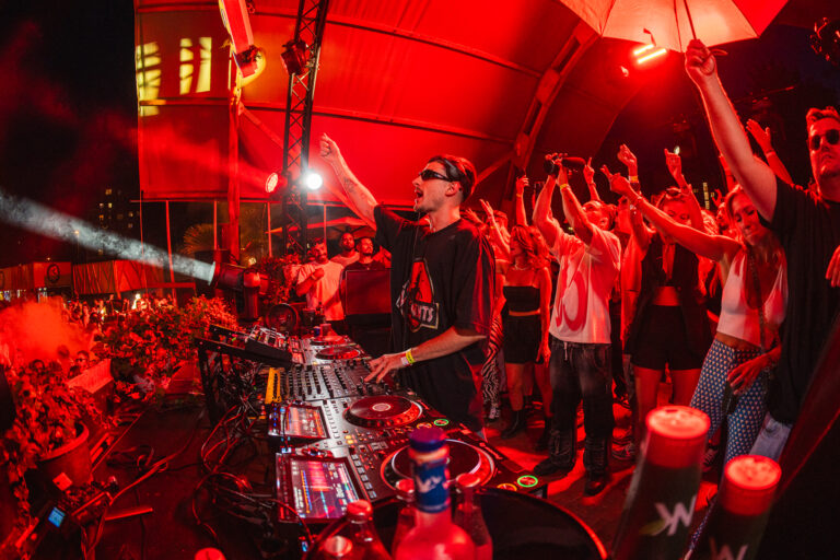 A man stands behind a mixing console under red stage lights, raising his fist as a crowd dances nearby.