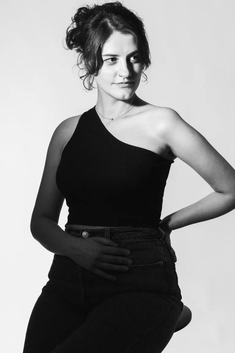 Black and white portrait of a woman wearing a one-shoulder top and jeans, posing against a light background.