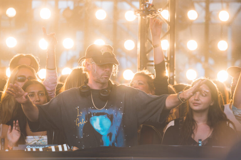 A man in a graphic t-shirt and cap stands with outstretched arms behind a mixing console, set against a backdrop of bright stage lights and a crowd.