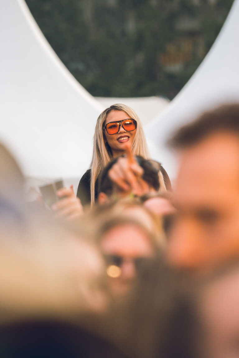A woman wearing orange tinted sunglasses stands out from the blurry crowd in natural outdoor lighting.