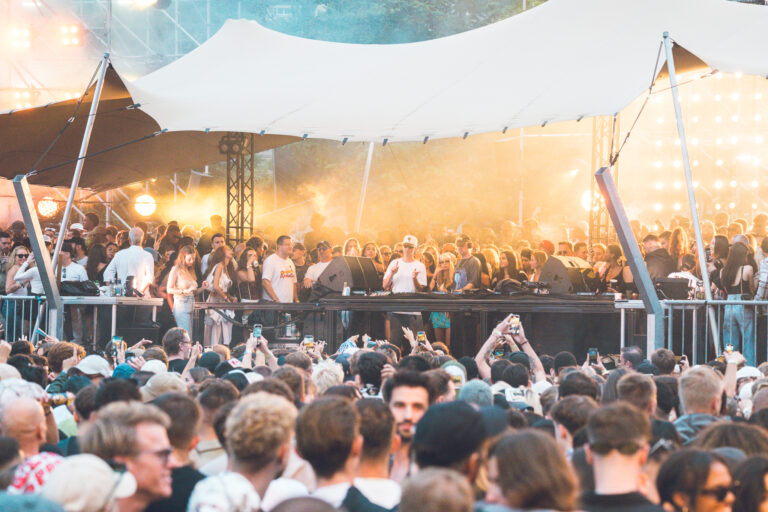 A large crowd gathers under a festival canopy in front of an elevated stage, with people holding up phones to capture the performance.
