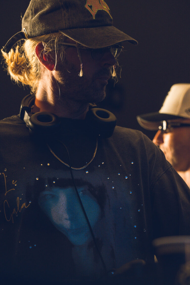 A man wearing a cap, glasses, and a dark printed t-shirt with headphones around his neck stands in warm lighting, with another person partially visible in the background.