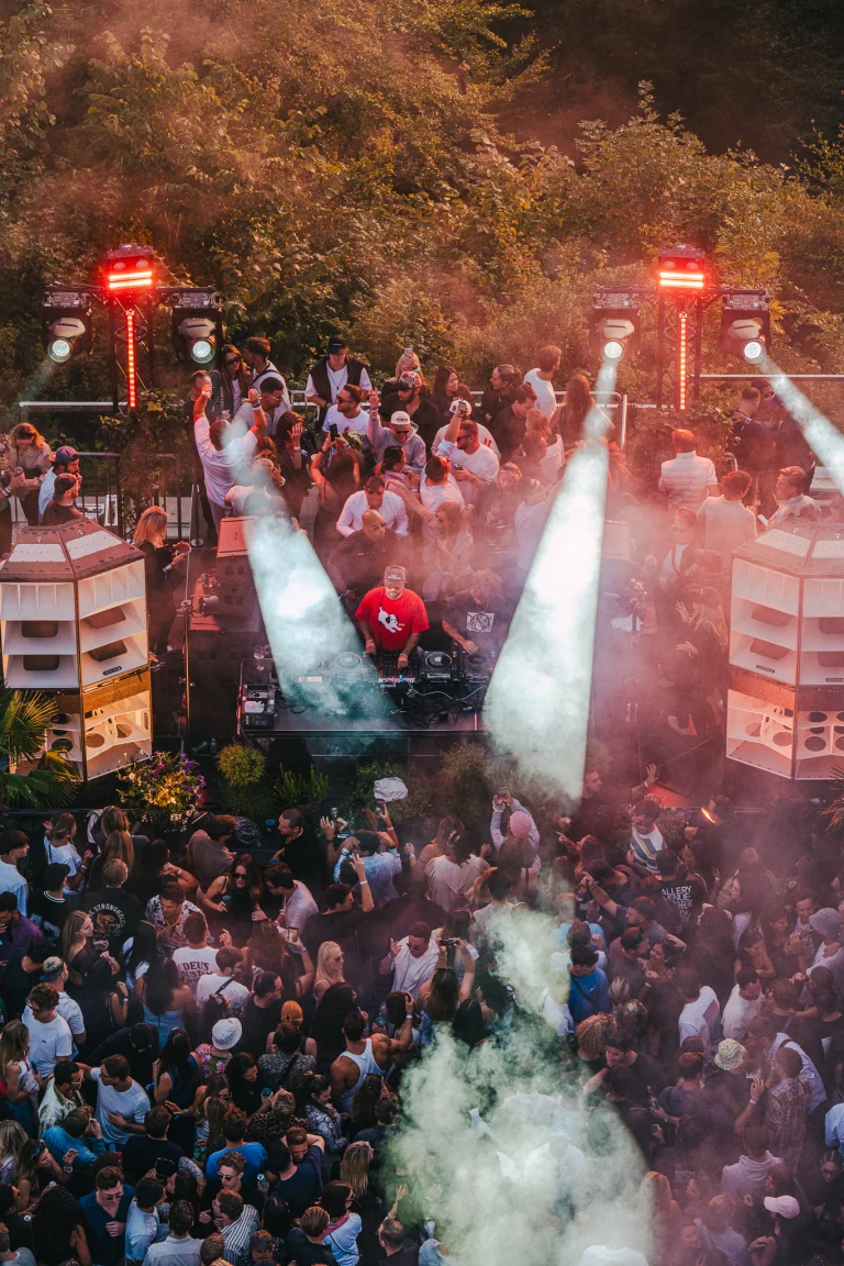 High-angle view of a crowded outdoor festival crowd facing a DJ booth flanked by large speaker stacks and stage lights cutting through smoke.