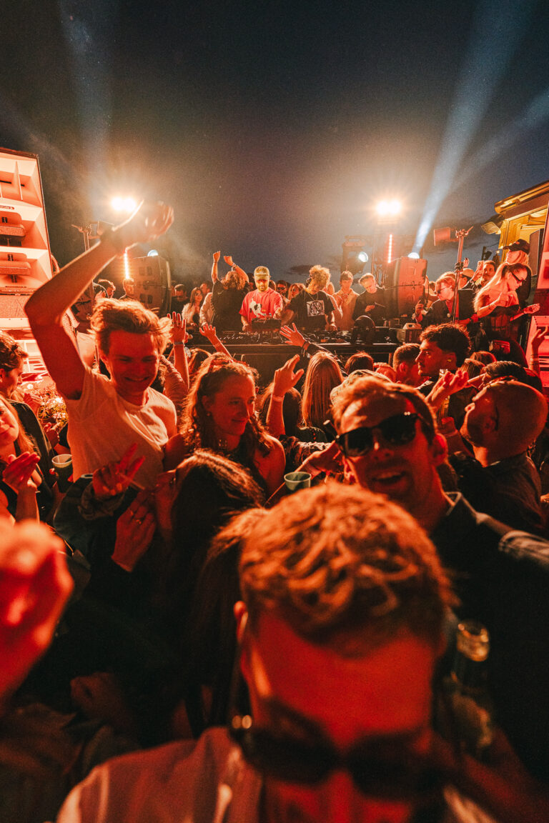 A crowd of people dancing outdoors at a festival under red lighting and stage lights at night.