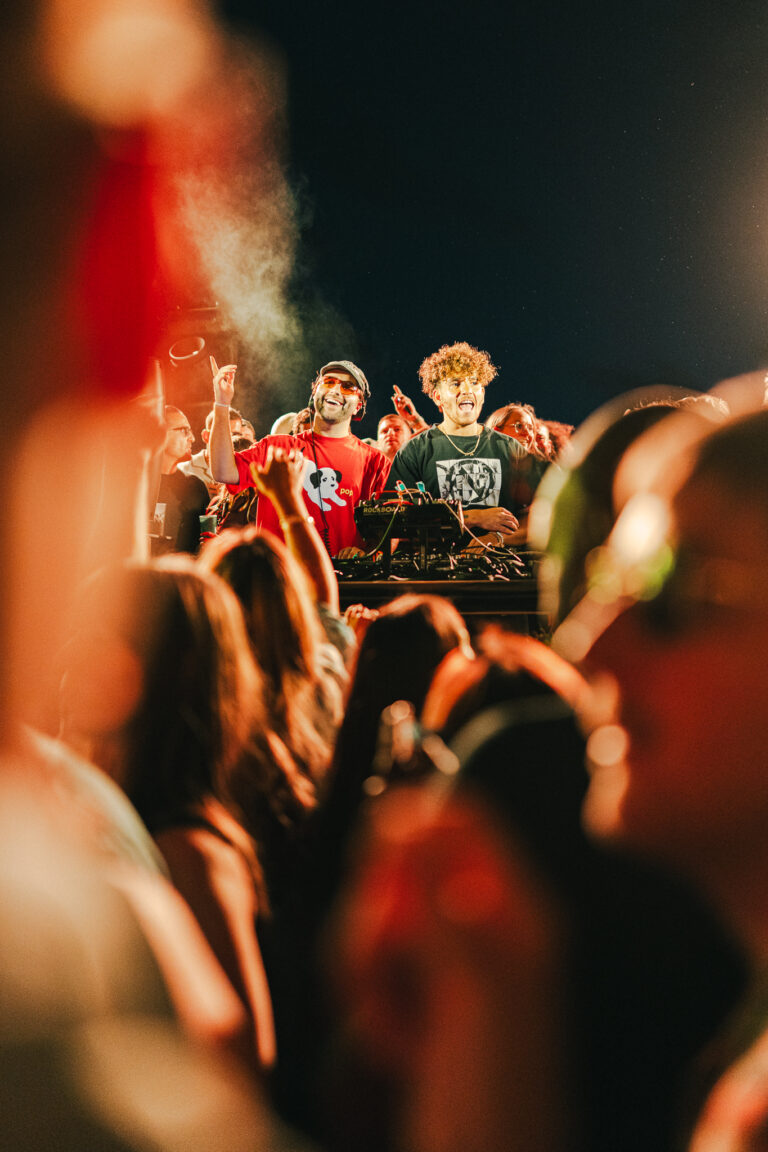 Two men behind a DJ booth perform at a festival under stage lights, framed by blurred silhouetted crowd members in the foreground.