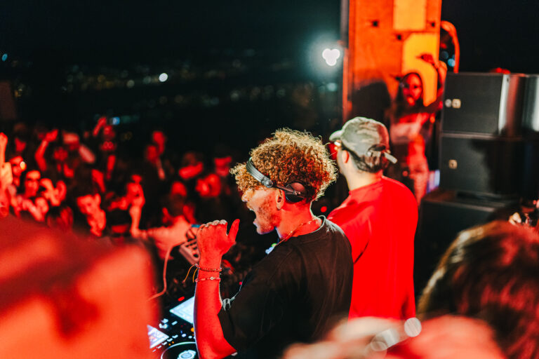 A man with curly hair wearing headphones stands at a DJ mixer before a large crowd under red stage lighting.
