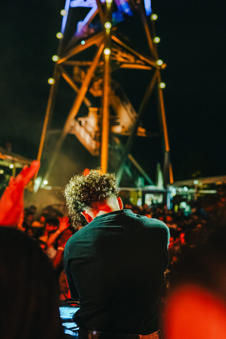 A man with curly hair stands in front of a tall, illuminated tower at night, facing away from the camera before a crowd.