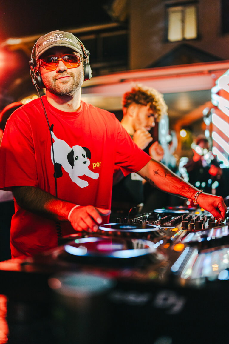 A man wearing tinted glasses and a red t-shirt with a dog graphic operates a DJ mixer under warm artificial lighting.