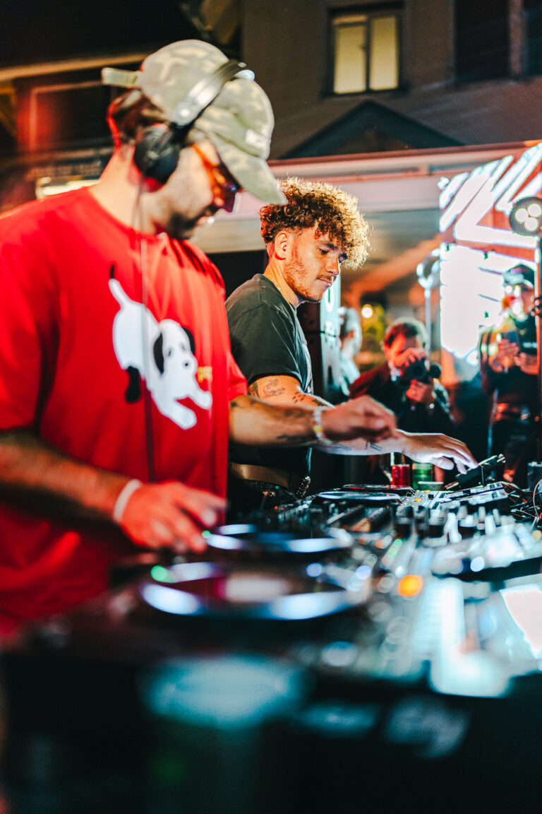 Two men standing behind a mixing console, with the man in the foreground wearing a red shirt featuring a white dog graphic and a cap.