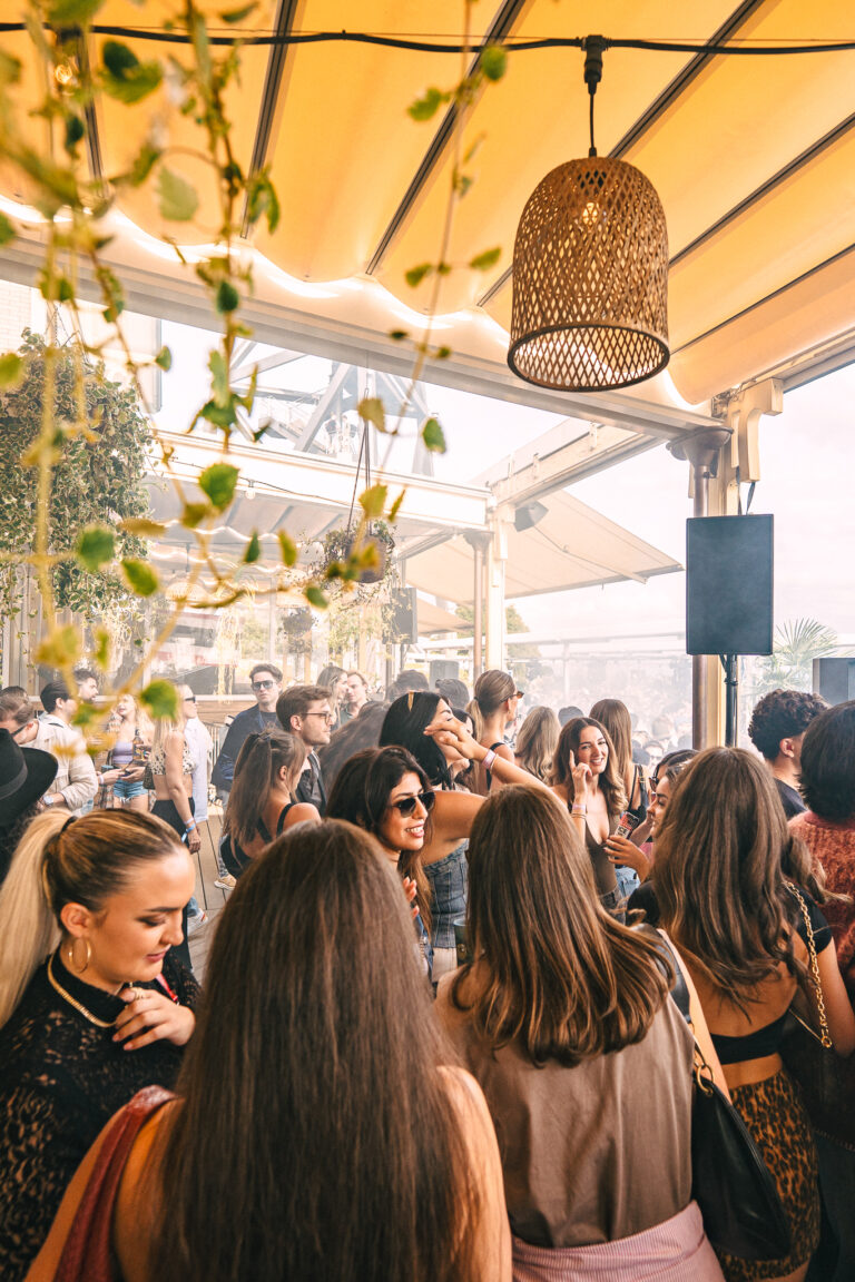 A crowded outdoor terrace filled with women and men socializing under a fabric canopy and woven pendant lamps.