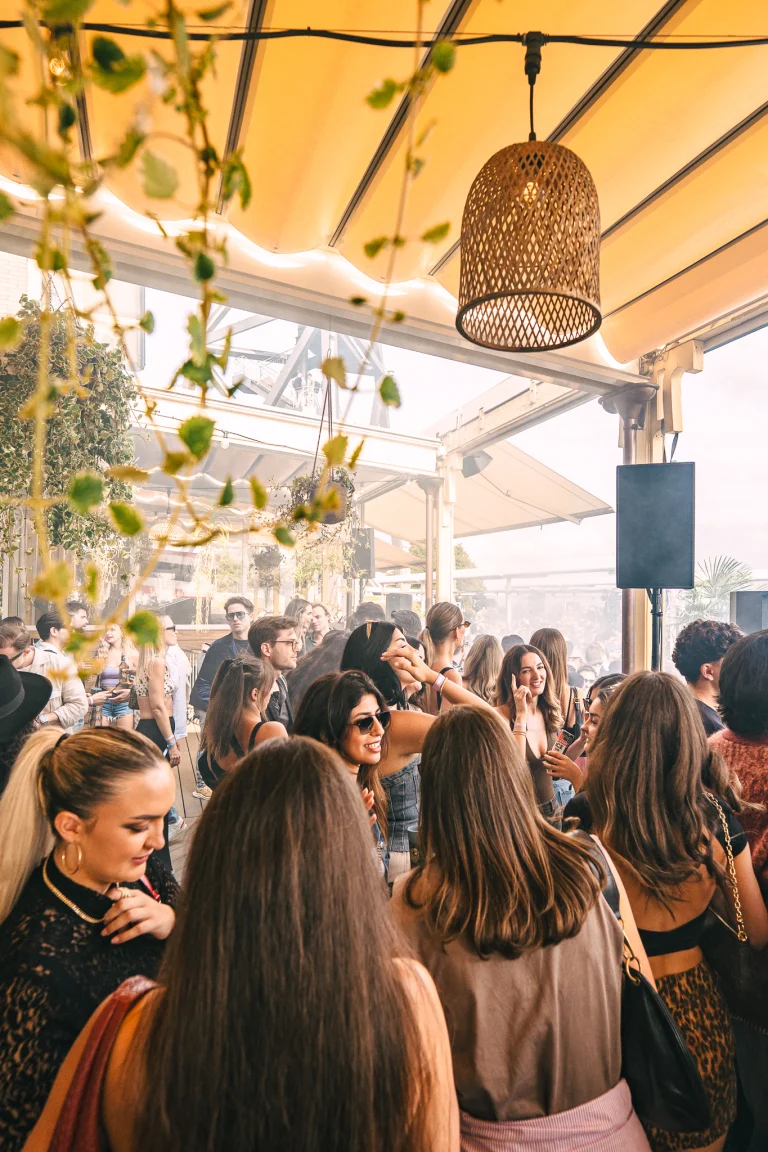 A crowded outdoor terrace filled with women and men socializing under a fabric canopy and woven pendant lamps.