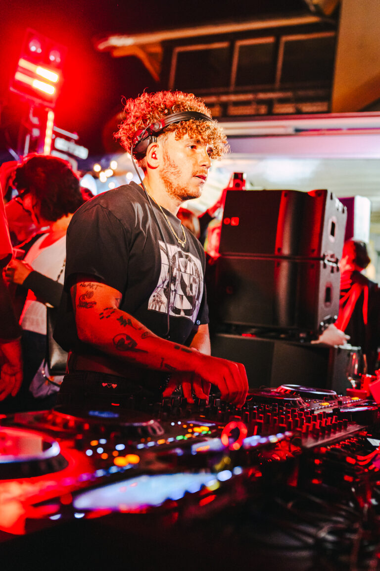 A man with curly hair and headphones around his neck stands behind a DJ mixing console under red stage lighting.