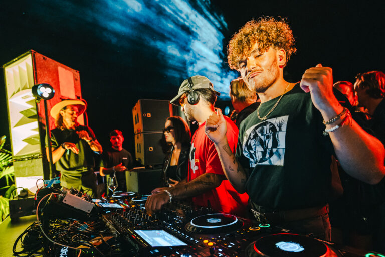 A man stands in the foreground at a DJ mixing console, with other people operating equipment in the background under nighttime stage lighting.