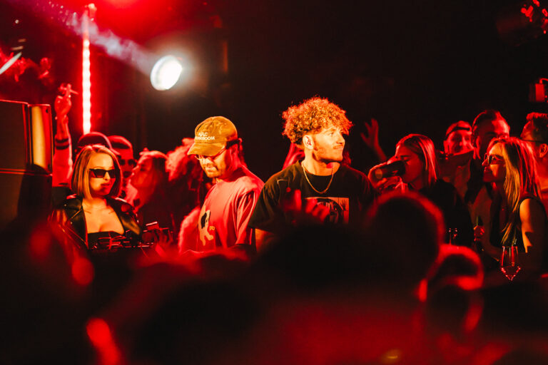 A dimly lit club filled with a crowd under red stage lighting, featuring a man with curly hair standing near the center among people.