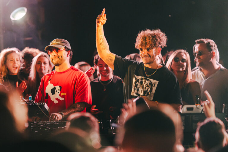 Two men standing behind a mixing console on a stage under warm stage lighting, with one man raising his arm amidst a crowd.