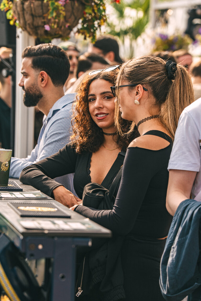 Two women stand at an outdoor bar in natural daylight, with a man visible in profile to their left.