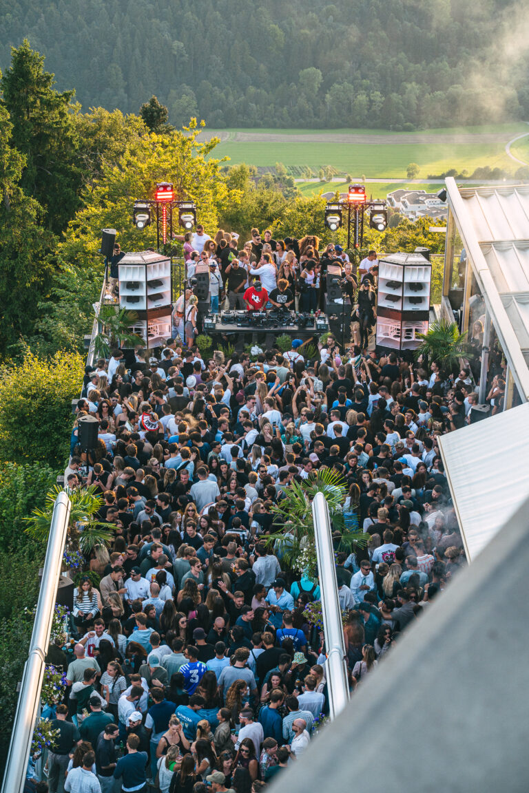 High-angle view of a large crowd gathered outdoors at a daytime festival, facing a raised DJ booth flanked by large speakers with a hilly landscape in the background.