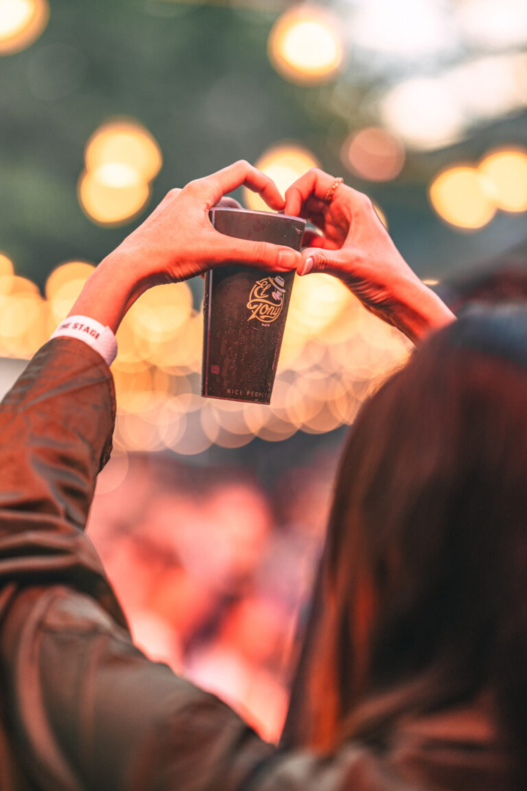A woman holds up a branded cup with both hands, shaping her fingers into a heart above it against a backdrop of warm bokeh lights.