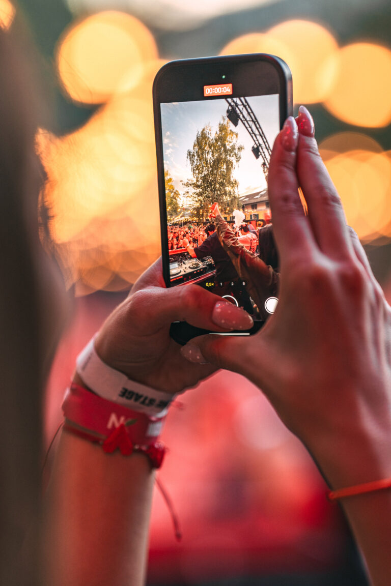 A woman holds a smartphone vertically, recording video of a festival stage and crowd displayed on the screen against a blurred background.