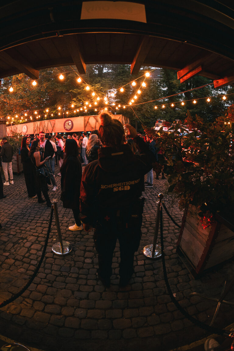 A woman in a security jacket stands on cobblestones, facing a crowded outdoor event lit by string lights under a fisheye lens.