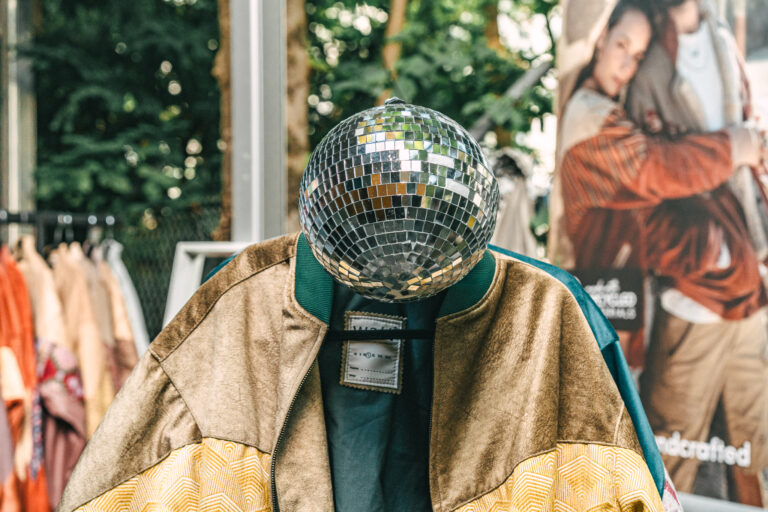 A disco ball hangs where a head would be above a jacket on a hanger, with clothing racks and a poster in the background.