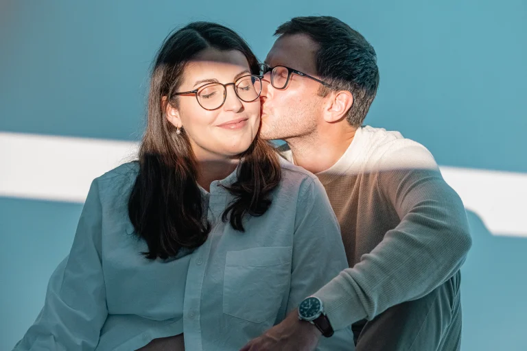A man wearing glasses kisses a woman wearing glasses on the cheek.