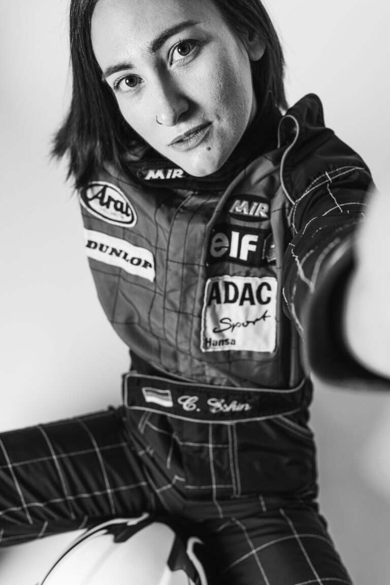 A black-and-white portrait of a woman wearing a racing suit with sponsor patches, looking down toward the camera.