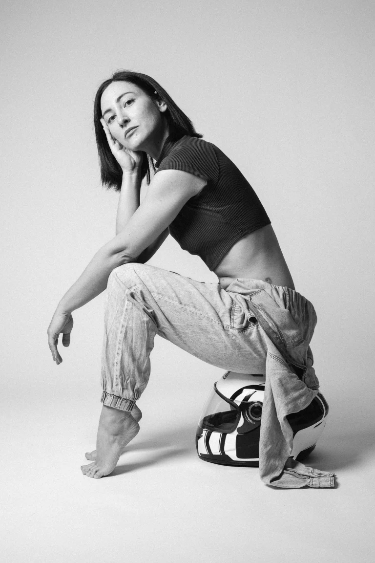 A black and white portrait of a woman crouching and balancing on a motorcycle helmet.